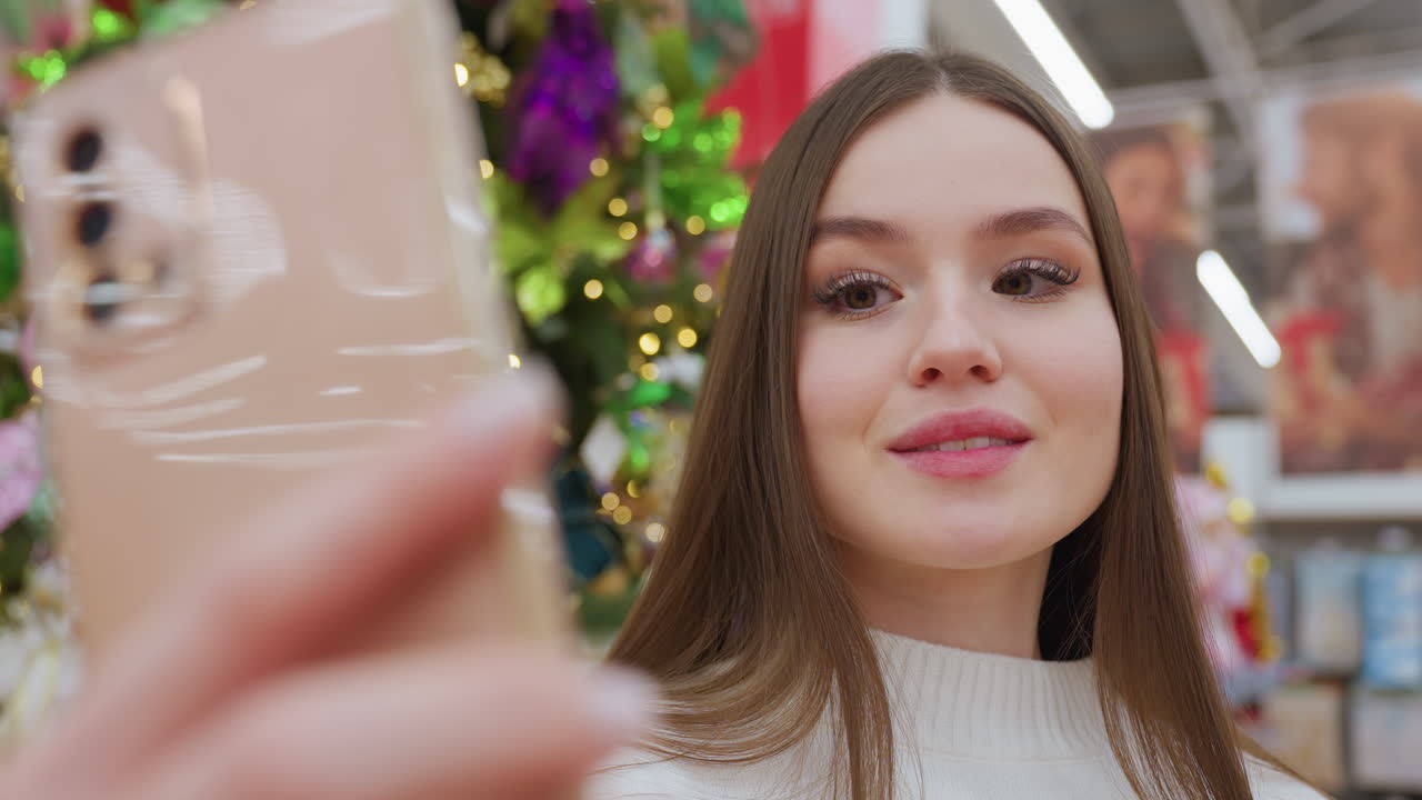 Close-up of a stylish lady taking a picture in front of vibrant Christmas decor in a well-lit store, she poses and blinks her eyes while smiling, surrounded by festive ornaments and glowing lights