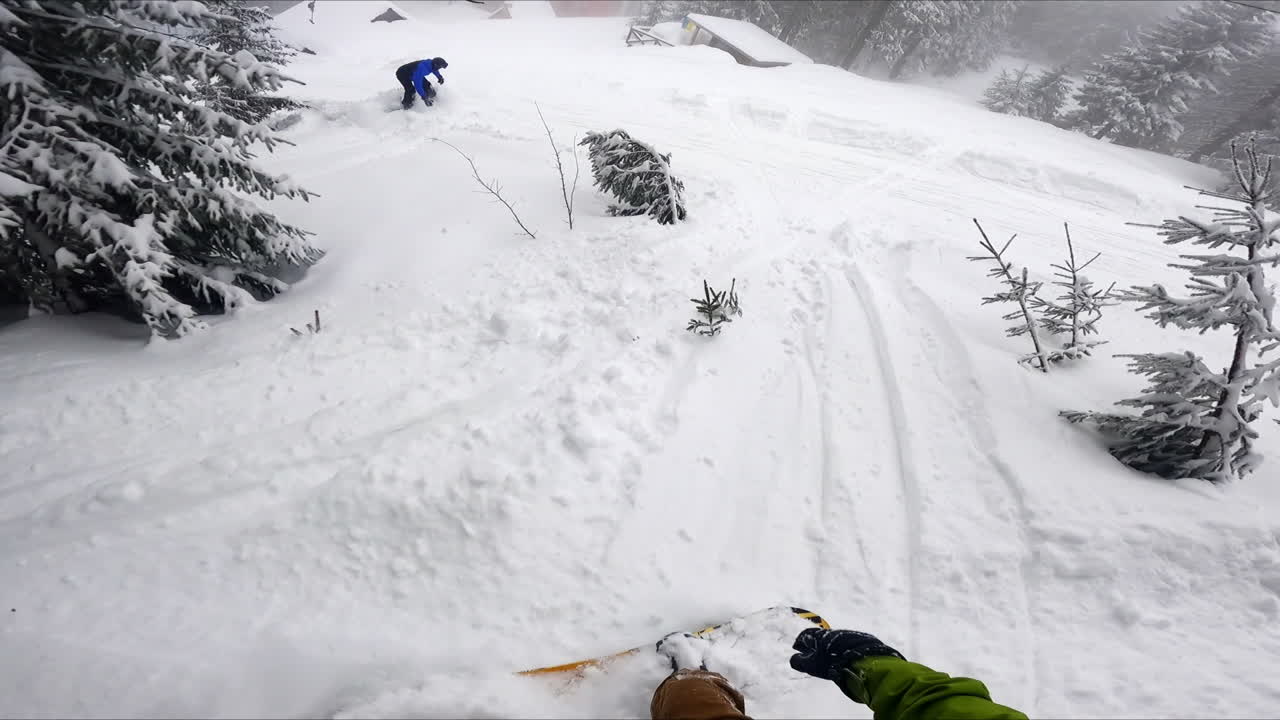 Two snowboarders move down the hills covered with thick layer of snow. Sportsmen arrive to the cottages.
