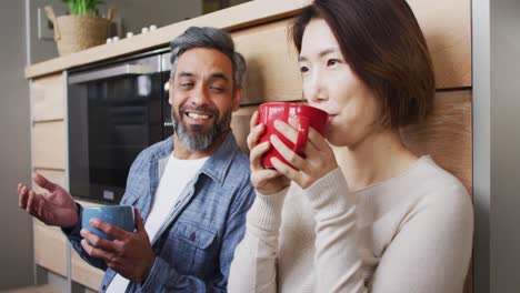 happy diverse couple sitting in kitchen drinking coffee and talking