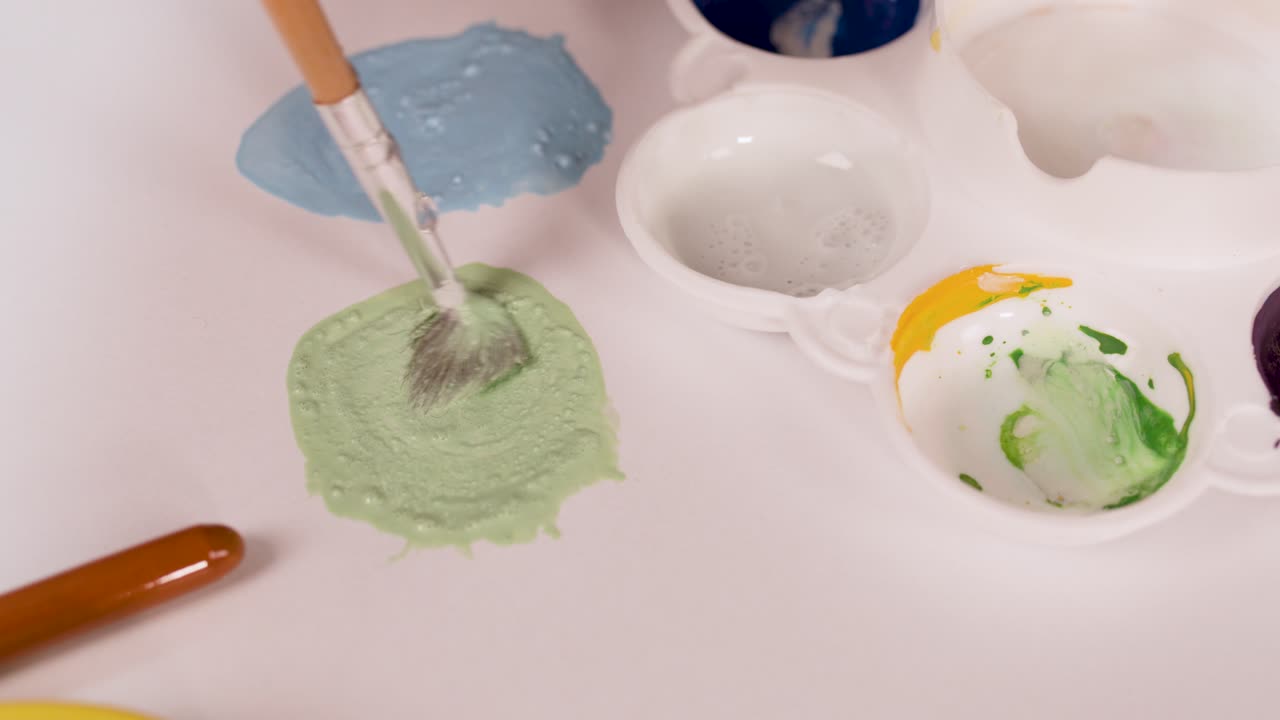 Hand mixes watercolor paints with brush on palette, surrounded by art supplies, under bright lighting