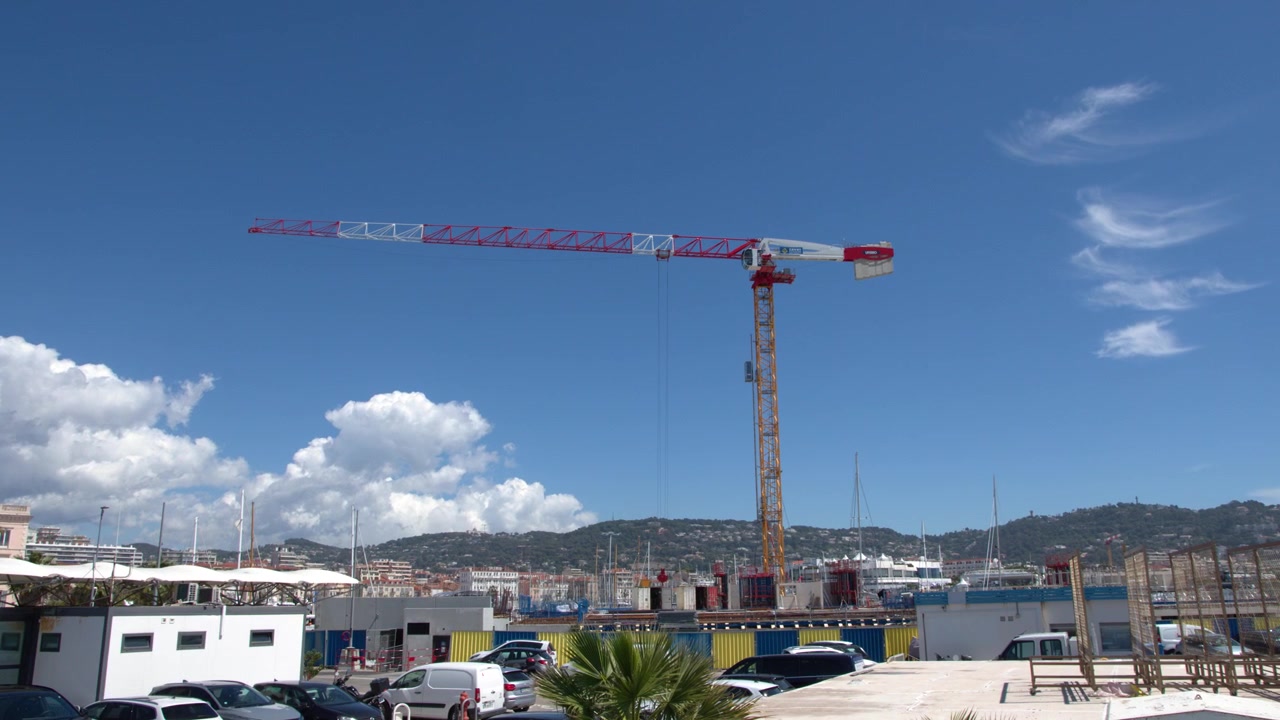 Inquadratura ampia di una gru rossa e bianca che ruota su un paesaggio urbano mediterraneo soleggiato