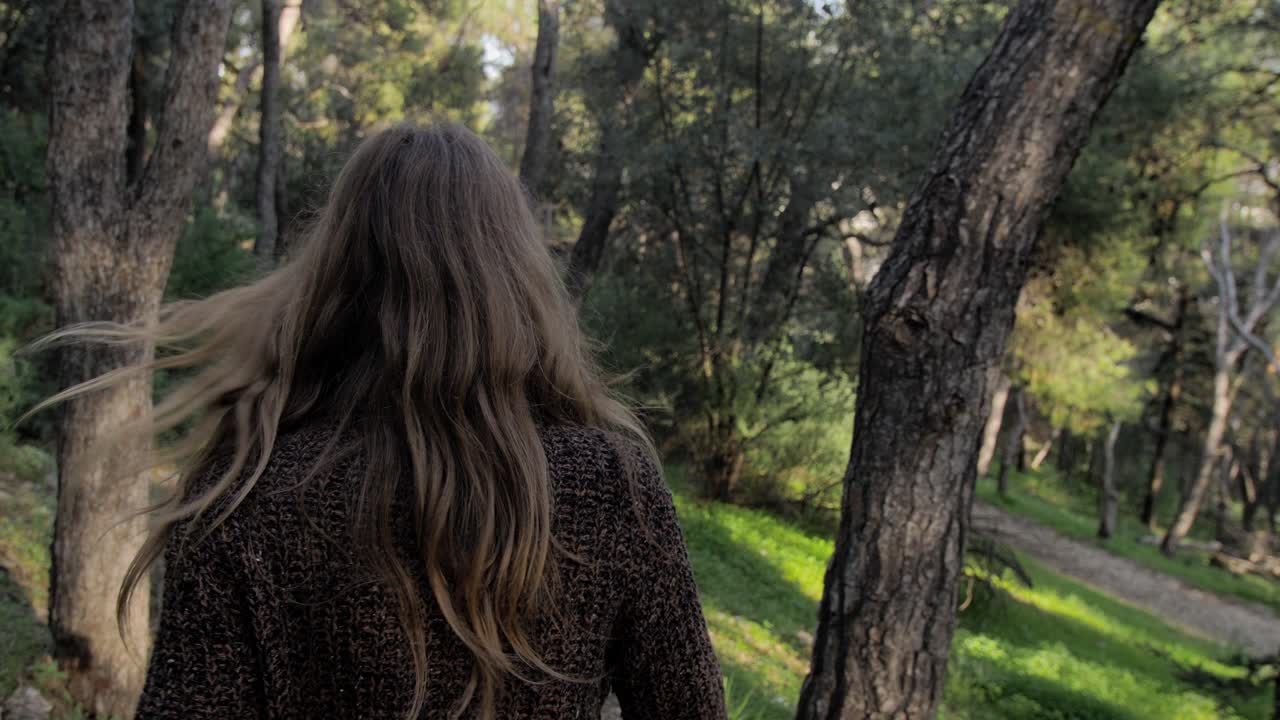 En cámara lenta, una mujer joven rubia camina por un sendero entre pinos, se gira para sonreír a la cámara, con seguimiento de gimbal