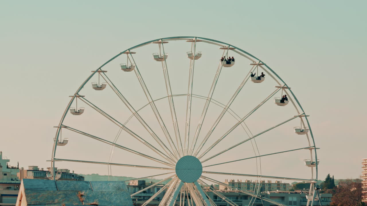 A wide view of a large Ferris wheel against a pale sky, with city rooftops visible below