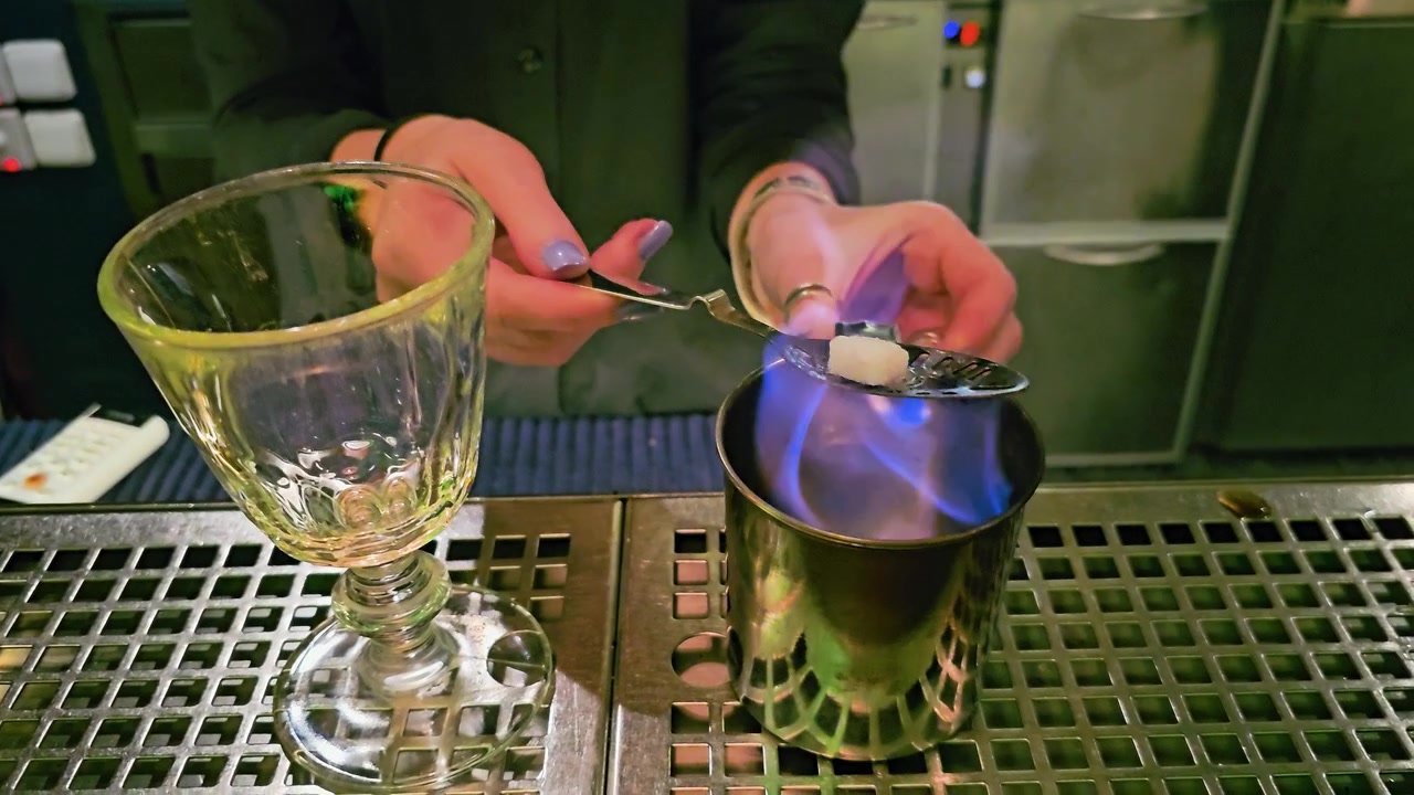 Bartender udfører klassisk absintritual med en flammende sukkerknald på en perforeret ske, metalblandekop, tomt glasbæger, grøn belysning i baren og stemningsfuld loungeatmosfære indendørs