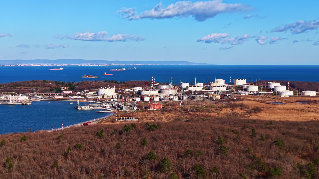 Vy över industriområde nära kusten med lagertankar och marina fartyg synliga på avstånd vid klart dagsljus och klar himmel.