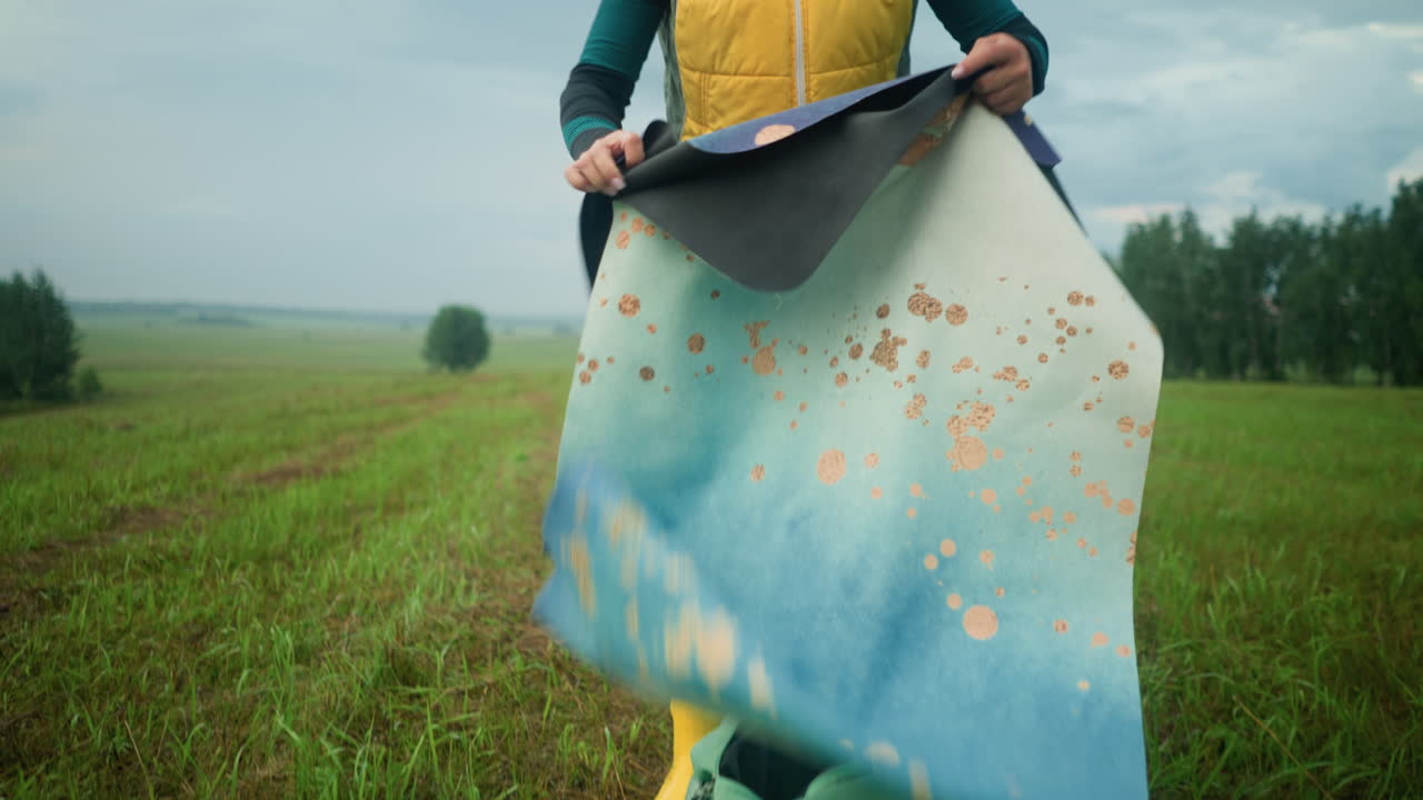 노란색과 초록색 재을 입은 여자가 잔디에 놓인 가방에서 매트를 가져옵니다. 그 에는 초록색 코트가 있습니다. 그녀는 머리카락을 조정하는 동안 매트를 집어 들고 광활한 야외에 집중합니다.