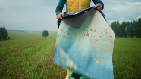 mujer en chaqueta amarilla y verde recupera alfombra de la bolsa colocada en el campo de hierba con abrigo verde a su lado, ella recoge alfombra mientras ajusta su cabello, mirada enfocada en el vasto campo abierto