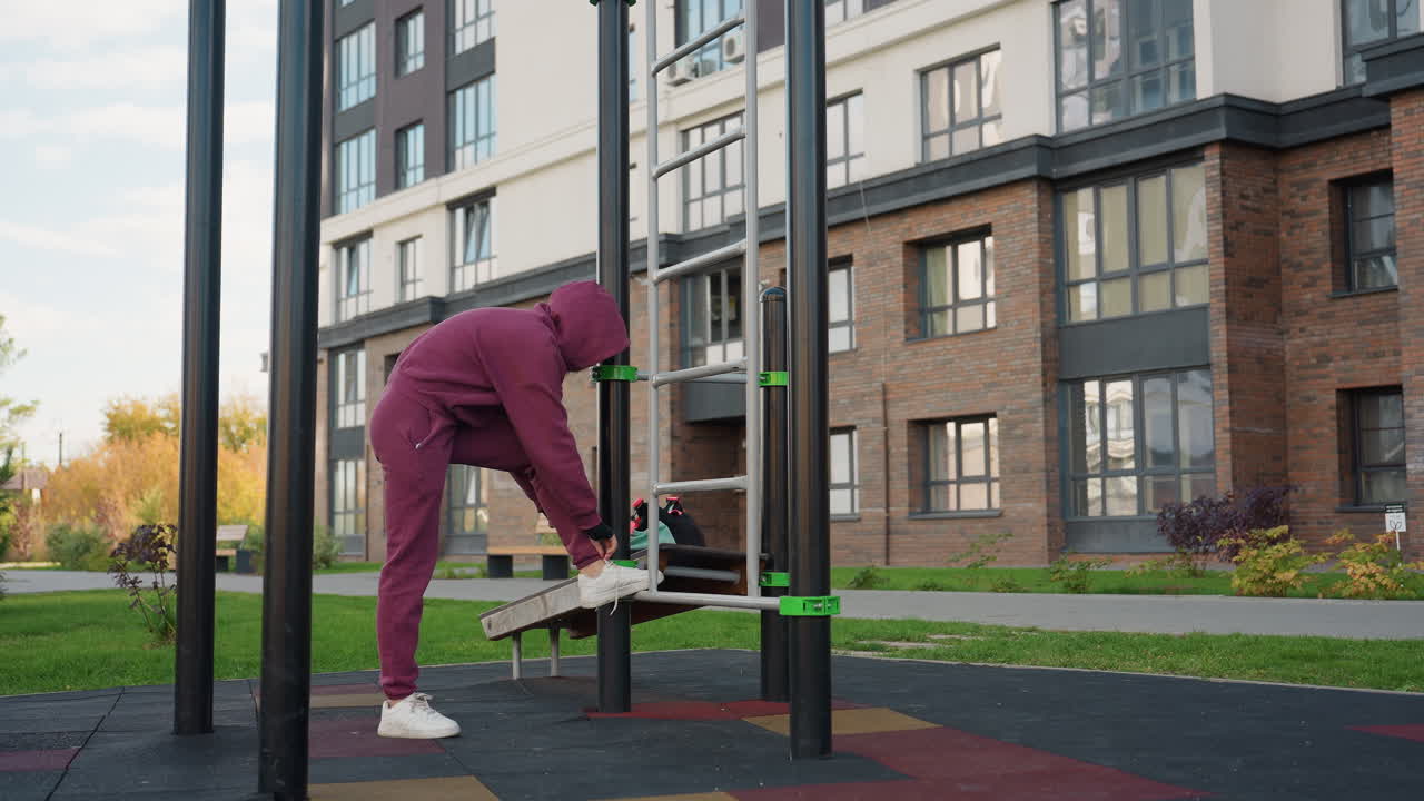 Casual sport woman walks toward outdoor bars carrying gym bag sets bag at corner then props leg on bar to tie shoelace for workout prep under clear daytime sky in urban park near modern building