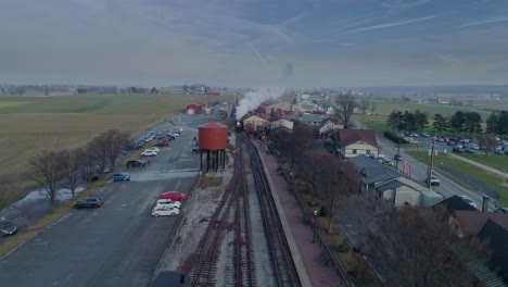 an aerial view of a train station, with a steam passenger train, pulling into the station, blowing smoke, in slow motion on a partially sunny day