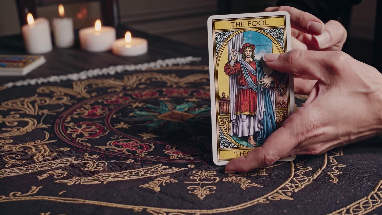 adivino místico colocando la carta del tonto del tarot en un ornamentado mantel negro y dorado, rodeado de velas iluminadas y rosarios, que simbolizan la guía espiritual y la predicción esotérica