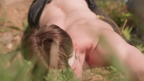 adventurer with head band crawls up rugged mountain slope under sunlit, tightly gripping grass with one hand for support, straining muscles visible, surrounded by green vegetation and rocky dirt path