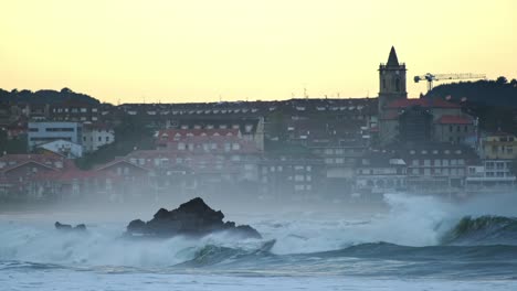 Grandes-Olas-Rompiendo-Sobre-Las-Rocas-Con-Los-Edificios-De-La-Ciudad-Al-Fondo-Por-La-Noche