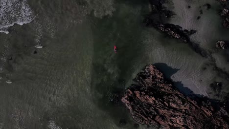 Person-on-a-paddle-board-heading-into-the-ocean-from-a-beach-with-rocks-and-waves