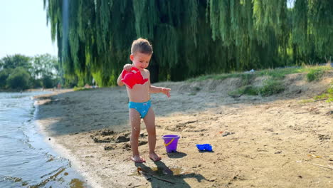 caucasian baby boy in blue swimming trunks plays at the river bank. smiling kid pours water from his watering can.