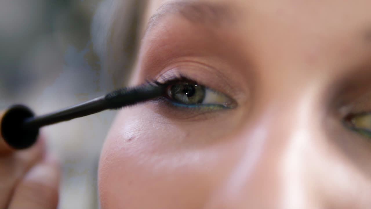 Extremely close of a young woman's eye. Make up artist carefully applying a black mascara. Caucasian model with green eyes