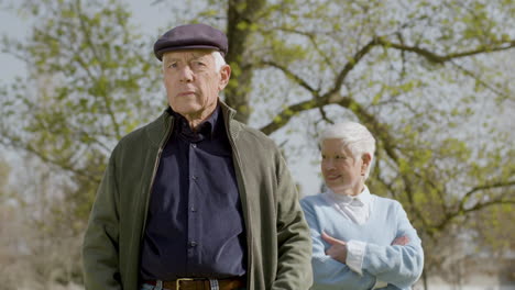 Portrait-Of-An-Elderly-Man-Looking-At-Camera-And-Smiling-While-His-Wife-Standing-Behind-On-A-Sunny-Autumn-Day-At-Park