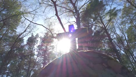 A-cross-in-the-sun-rays-on-the-top-of-a-stone-chapel,-with-a-pine-forest-in-the-background