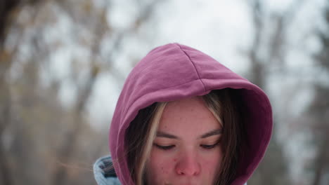 close-up of woman jumping outdoors in winter, wearing a hooded jacket, she has a slightly flushed face from the cold, against a blurred snowy background of trees and nature