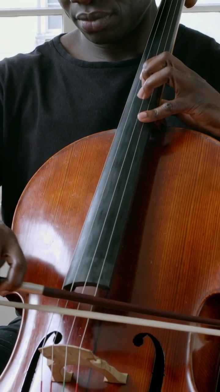 Vertical video: Starting bow stroke cellist wearing black tee practicing cello on neck by window