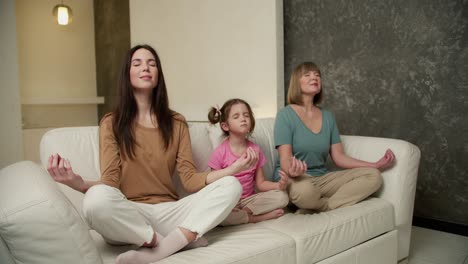 two mature women and a little girl are sitting on the sofa in a yoga pose and meditating