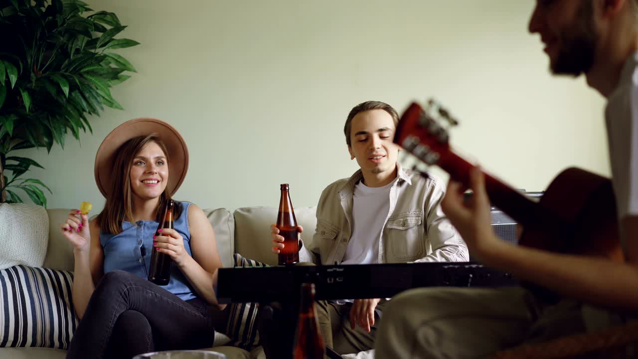 музыкальная группа делает перерыв во время репетиции, молодые люди разговаривают, едят закуски и пьют пиво. бородатый парень играет на гитаре, другие поют и смеются.