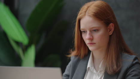 A-young-red-haired-business-woman-looks-thoughtfully-at-the-screen-and-brainstorms.-Watch-and-think-about-problems-looking-out-the-window.-Thoughtful-business-woman