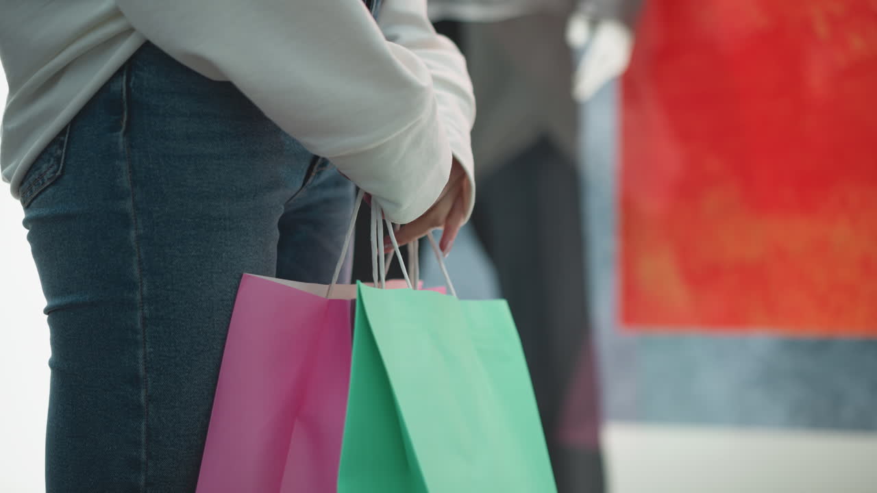 vista lateral de una mujer de pie en el interior sosteniendo bolsas de compras de menta y rosa, haciendo movimientos de la mano alrededor de las manijas con uñas pulidas, usando una camiseta blanca casual y vaqueros, fondo rojo y azul vibrante
