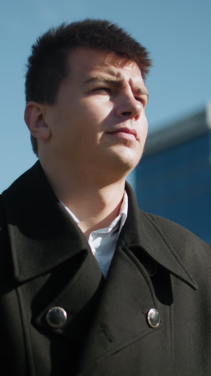 검은 코트를 입은 남자가 은 날 자신감 있게 산책하고 있습니다. 활기찬 파란 하늘 아래 배경에 흐릿한 건물과 나무가 있는 도시 건축물로 둘러싸여 있습니다.