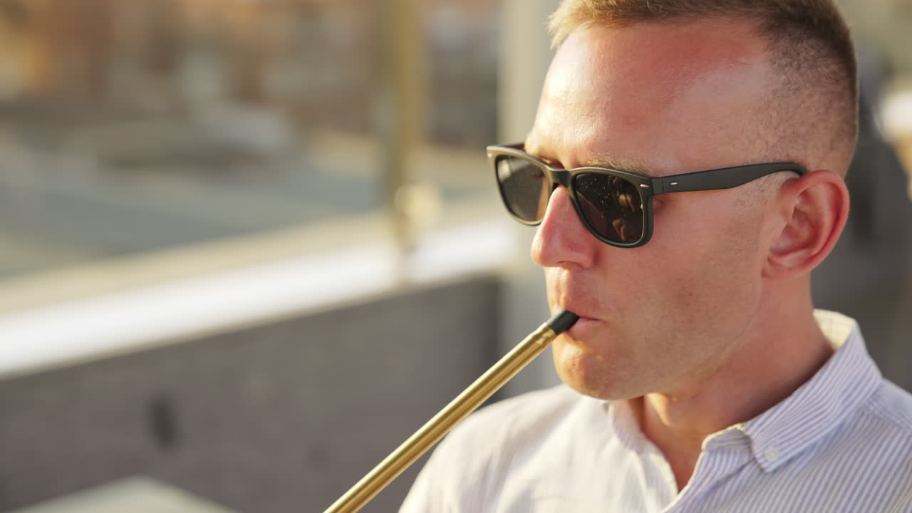Mid-aged man in sunglasses smoking chillum sitting outdoors. Chilling male rises head and breathes out white smoke. Close up.