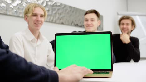 close-up of laptop with green screen with blurred men talking and smiling at background sitting in office conference room. chromakey device with webpage and coworkers discussing startup