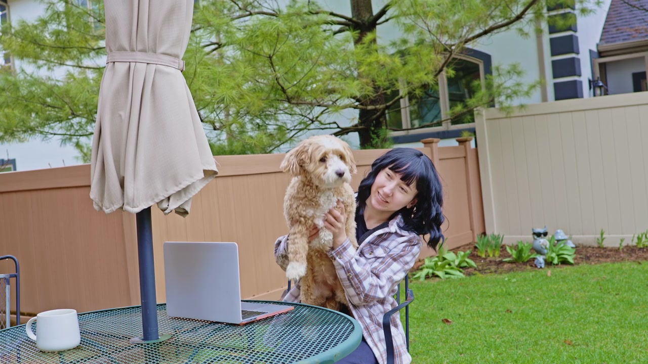 日中、裏庭で犬を抱きながら屋外でノートパソコン作業をする女性。
