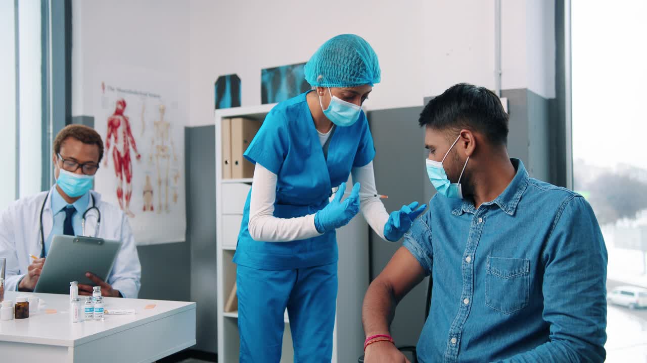 enfermera hindú muy joven o asistente de virólogo inyectando a un apuesto paciente masculino sentado en el gabinete del hospital con máscara médica mientras el médico trabaja y lee análisis, vacunación contra el coronavirus