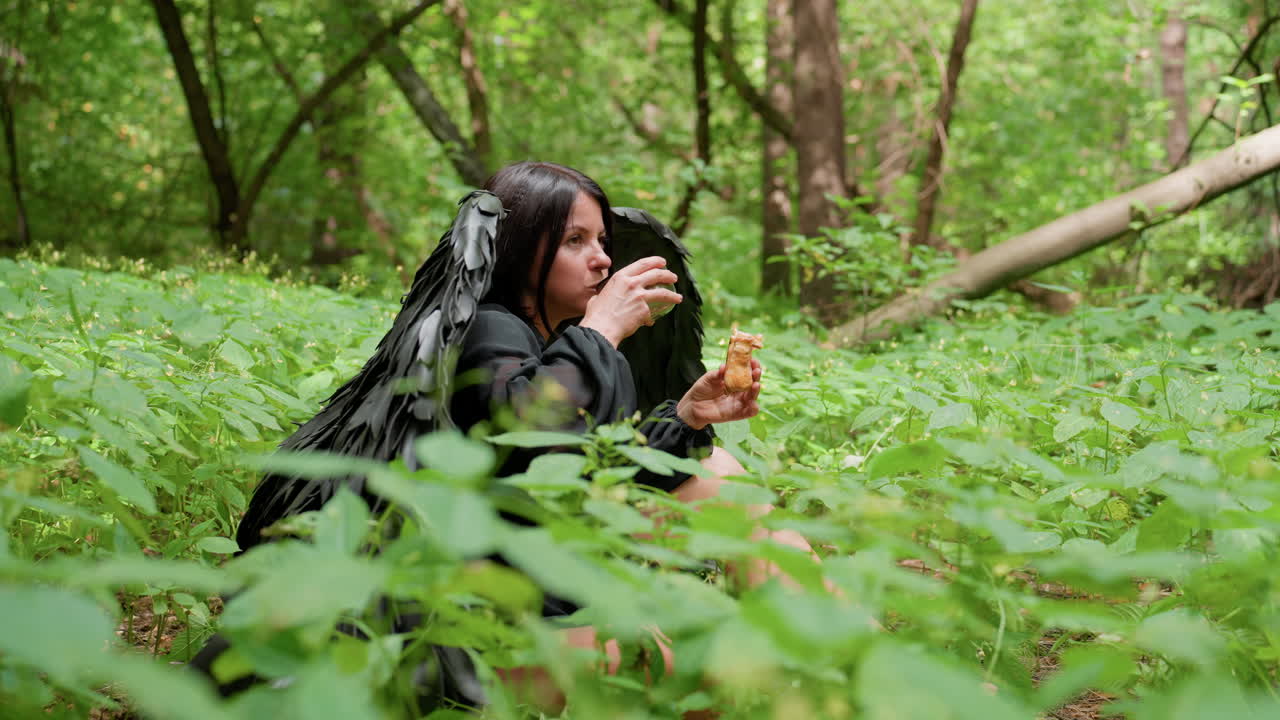 Side view of woman dressed in black winged costume sitting among green plants, chewing snack thoughtfully while sipping tea in peaceful forest setting under soft sunlight