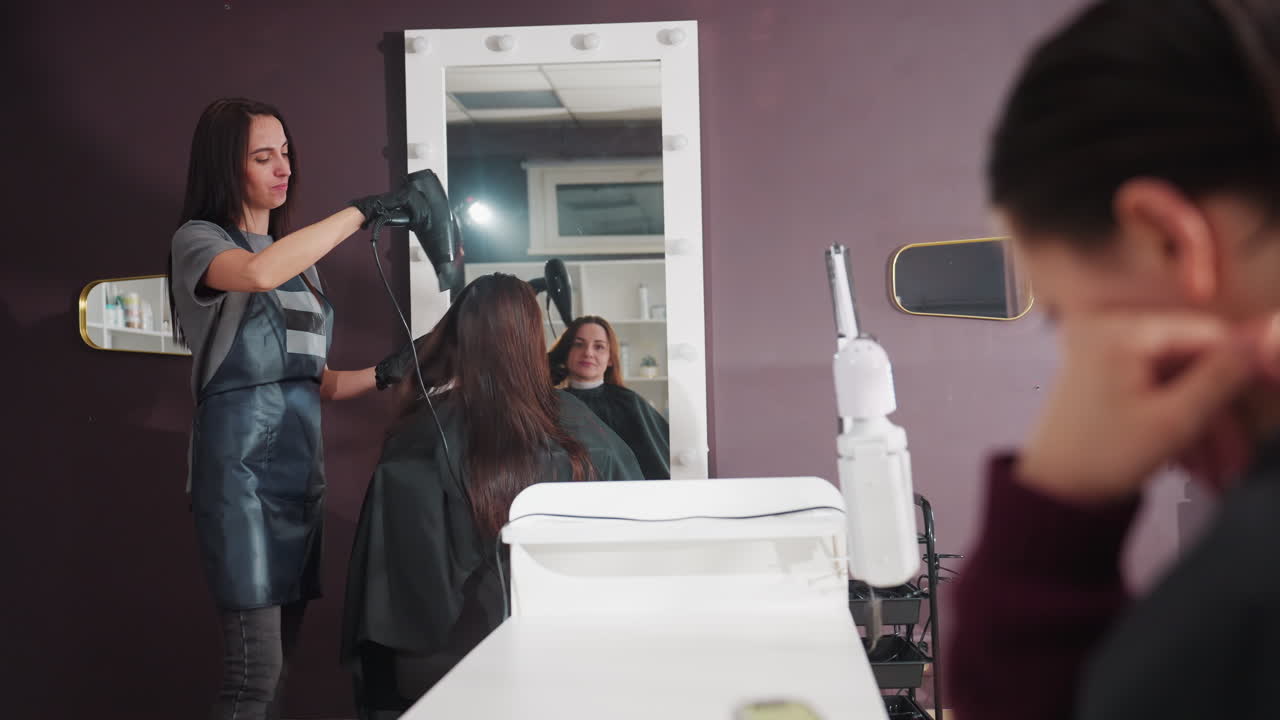 stylist drying client's hair in salon mirror, caucasian professional attentively blowouts and consults, stressed companion in foreground with hand on face, warm studio lighting, modern shelves