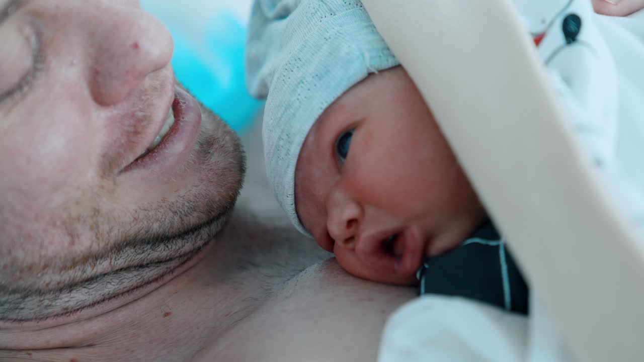 Loving dad kisses tenderly the head of his newborn child. Adorable baby winks and opens mouth. Close up.