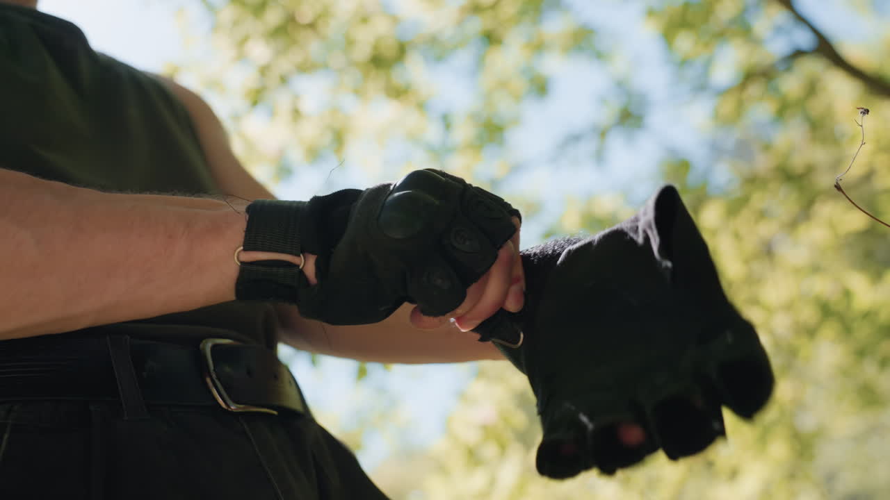 Under view of adventurer putting on tactical gloves and folding arms in readiness for next move, sunlight streaming through green leafy canopy overhead, wearing green cargo pants and belt