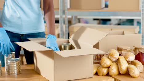 close-up view of african american volunteer hands packing box with cans of preserves in charity warehouse