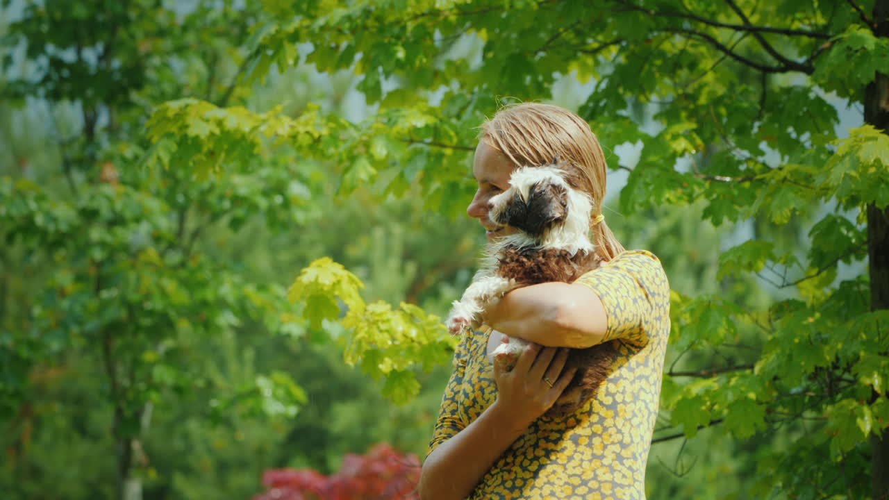 행복한 중년 여성은 비가 내리는 잊을 수 없는 순간에 정원에 서서 젖은 강아지를 껴안습니다