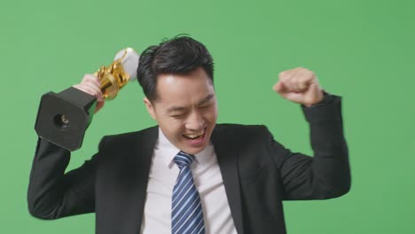 close up of asian business man in a suit and tie with a gold medal looking at a gold trophy then celebrating winning as the first winner on green screen background in the studio