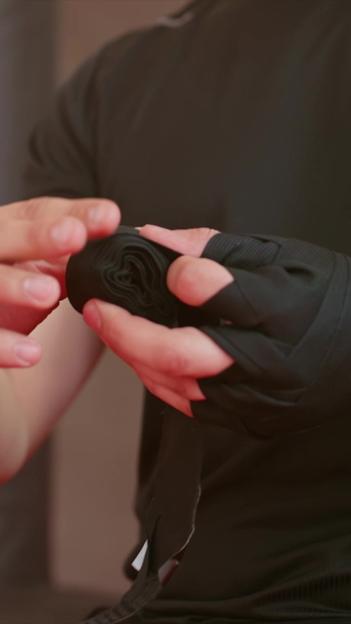 Man carefully wraps his hand with black elastic material. Close up view shows precise manipulation. Indoor setting with neutral background emphasizes focused action