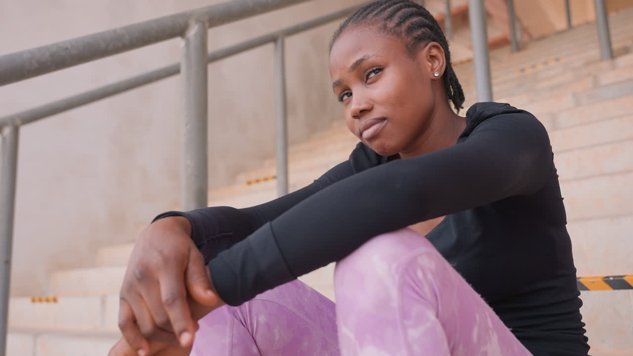black student sitting on steps, thoughtful gaze toward horizon casual athletic wear with purple leggings, braided hair, metal railing and concrete stairs, hands clasped over knees, quiet ambition