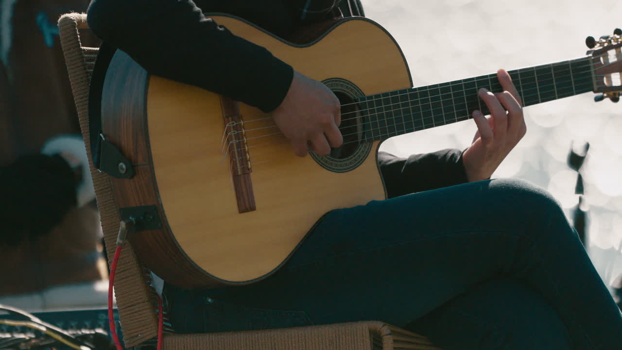 Detailed shot captures seated musician strumming classical guitar with both hands visible on fretboard and body, jeans and black top worn, soft background blur, real time, static shot