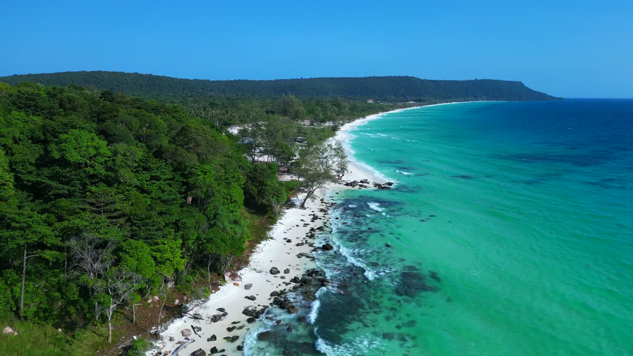 Black Stones in turquoise water gently washing the white sand beach of Koh Rong island, cambodia, on a sunny day with clear blue sky. Dramatic aerial view flight static tripod hovering drone