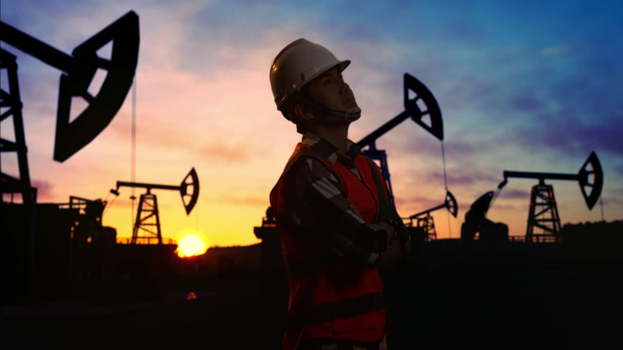 vista lateral de un ingeniero masculino asiático con casco de seguridad cruzando los brazos y mirando a su alrededor mientras está de pie frente a las bombas de petróleo, durante la puesta o salida del sol