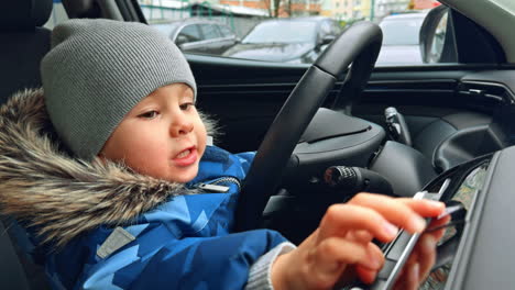 cute caucasian toddler in grey cap and jacket sits in a car. happy baby pressing buttons and moves the steering wheel. close up.
