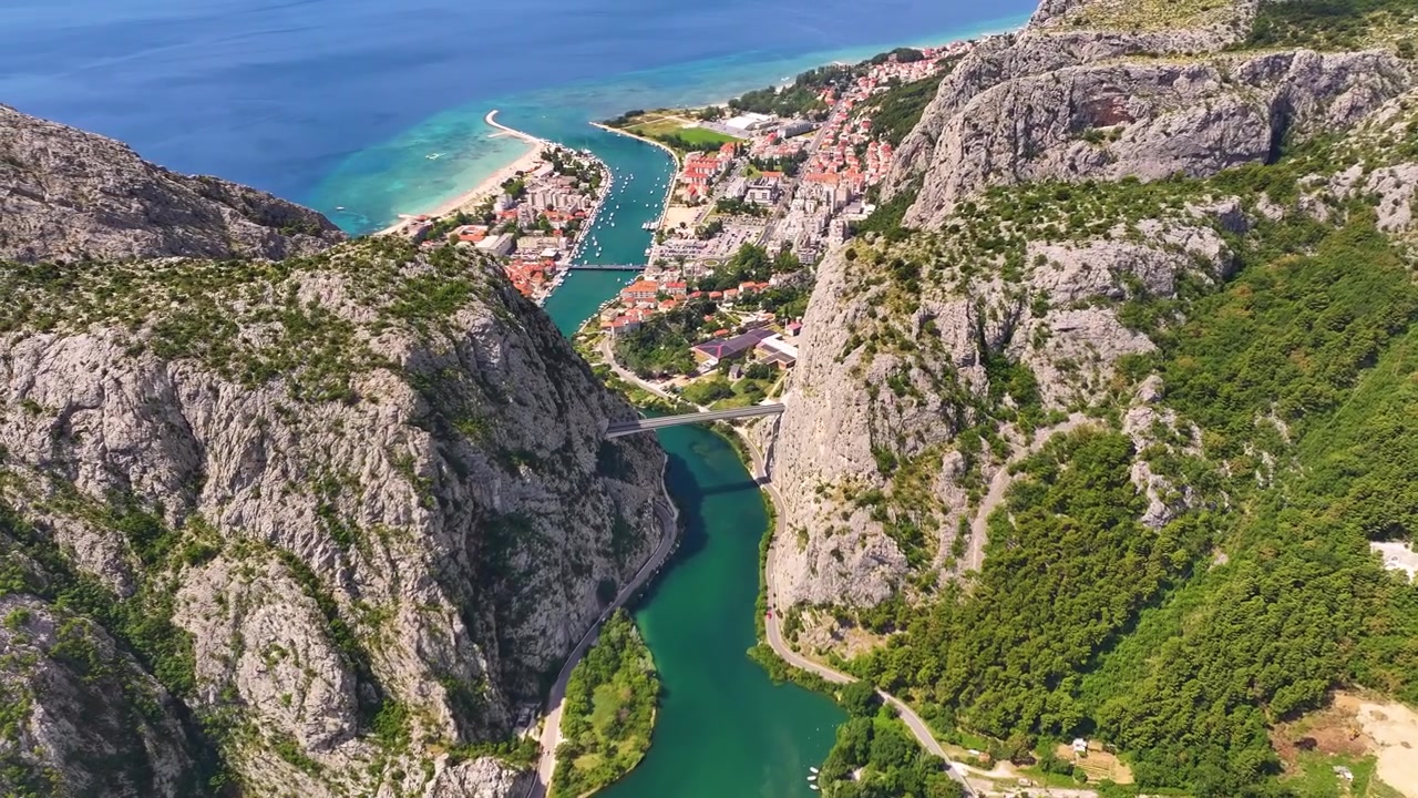 Letecký pohled na Omiš v Chorvatsku, ukazující řeku, nedaleké hory a pobřežní oblast. Město má několik budov, lodě ve vodě a most přes řeku.
