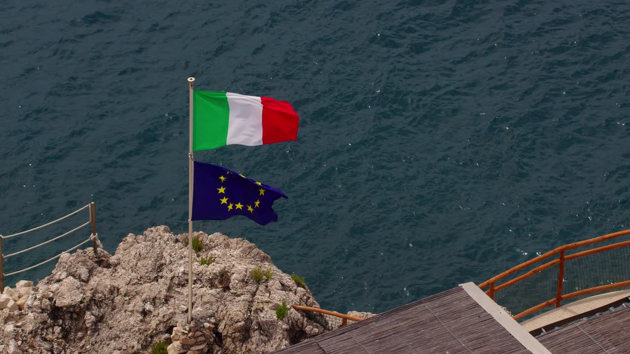 Italian and EU flags waving powerfully on rocky coast by the sea in Taormina, Sicily, Italy (Italia)