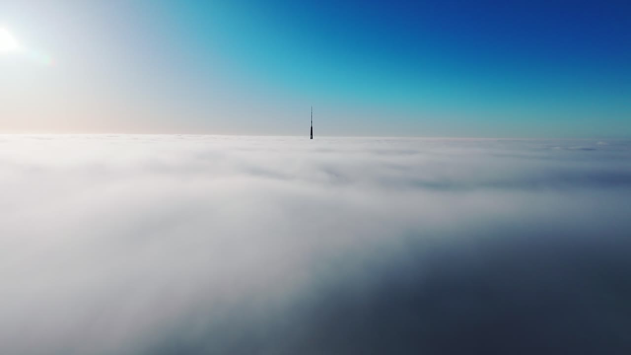 Forward drone movement above thick fog reveals distant Riga TV tower at sunrise
