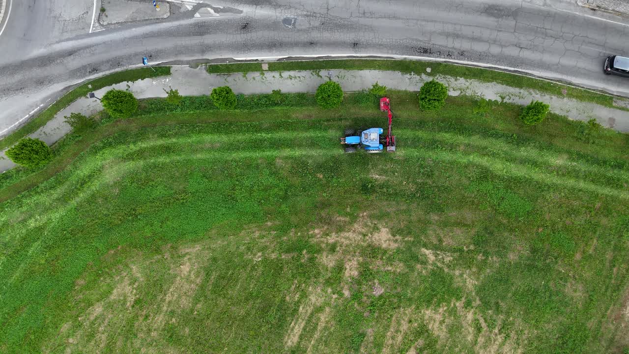 Aerial view of a farmer mowing grass with a tractor along a residential street in a suburban area of Piacenza, Italy, showcasing urban and rural coexistence