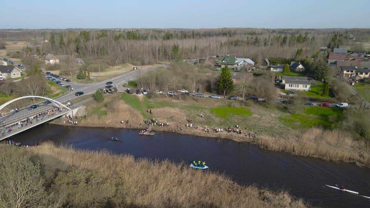 Aerial drone footage of people kayaking and paddle boarding with canoes and rowing on dark brown murky muddy river water underneath a bridge during a sunny day at Võhandu marathon. Spectators on shore